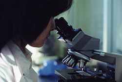 Person looking into a microscope in a lab setting. She has dark hair and wears a lab coat.