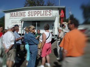 People gathered outside a marine supplies shop on a sunny day, with a red shop sign and palm trees in back.