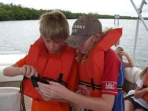 Two boys on a boat, wearing orange life vests, examining a small device.
