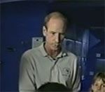 Man in a light-colored polo shirt looking down, possibly holding something dark. Blue background, dimly lit.