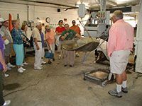 Group of people gathered around a large metal machine in a workshop, watching a demonstration.
