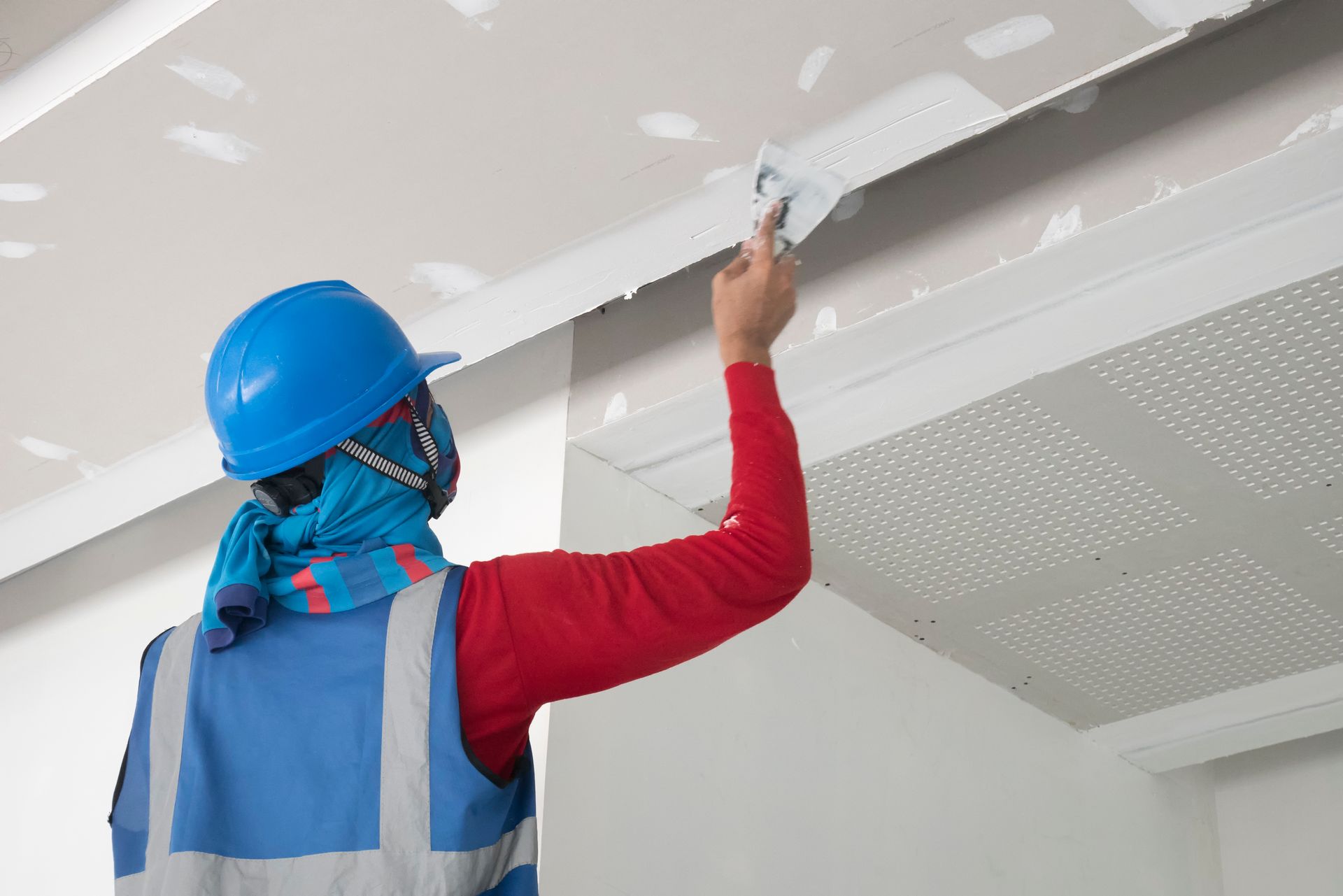 Person in blue coveralls and yellow hard hat painting a wall white with a roller.