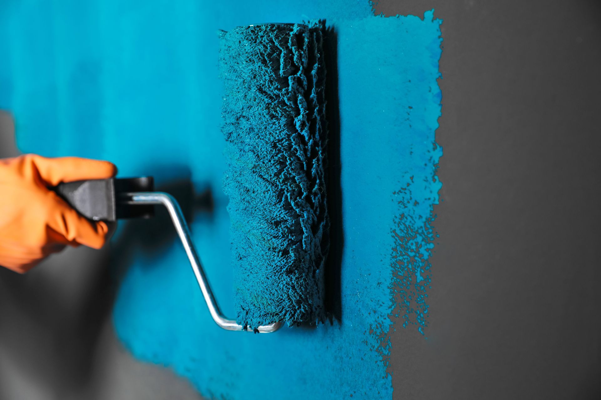 A gloved hand uses a paint roller to apply blue paint to a gray wall.