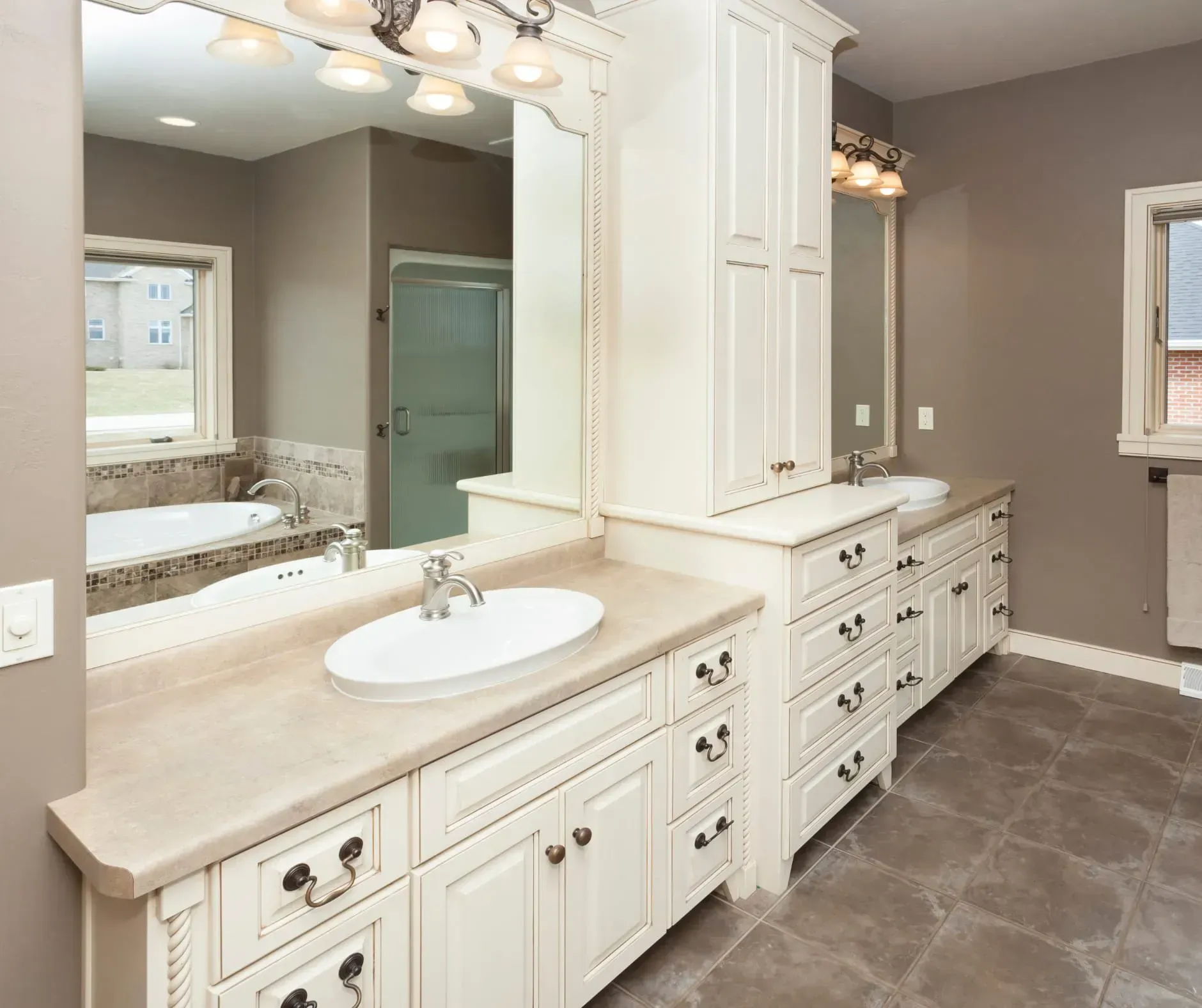 Bathroom with cream-colored cabinetry, dual sinks, large mirror, and a bathtub. Gray walls and brown tile floor.