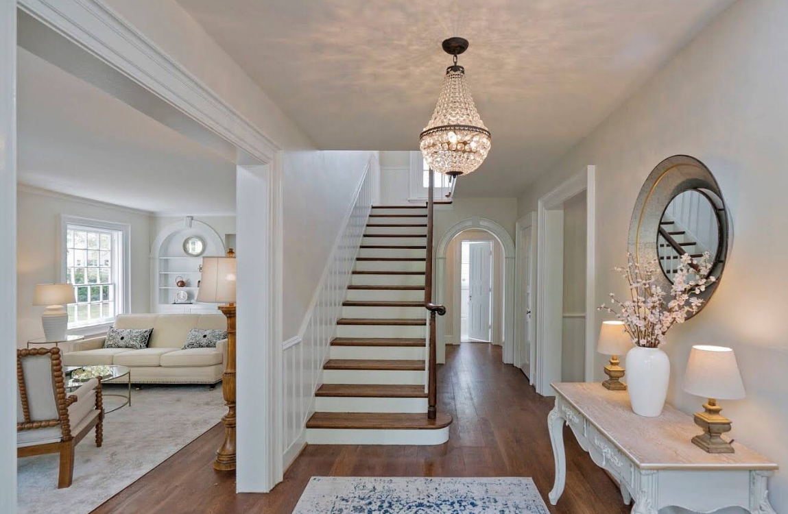 Bright hallway with staircase, chandelier, console table, and adjoining bedroom visible through an open doorway