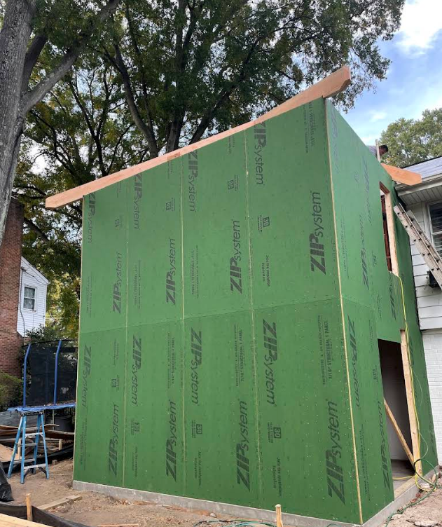 Green sheathed shed under construction beside trees, with slanted roof framing and an open doorway.