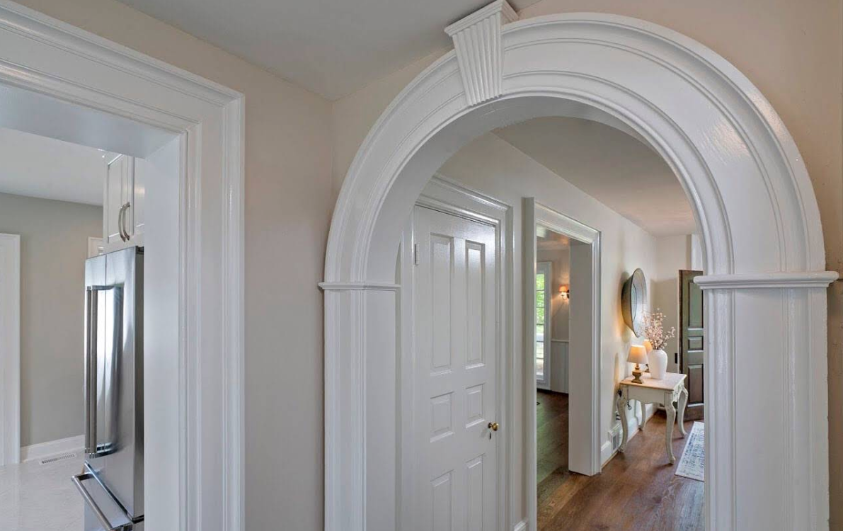Arched hallway with white trim, open doors, and a glimpse into a bright, furnished room beyond