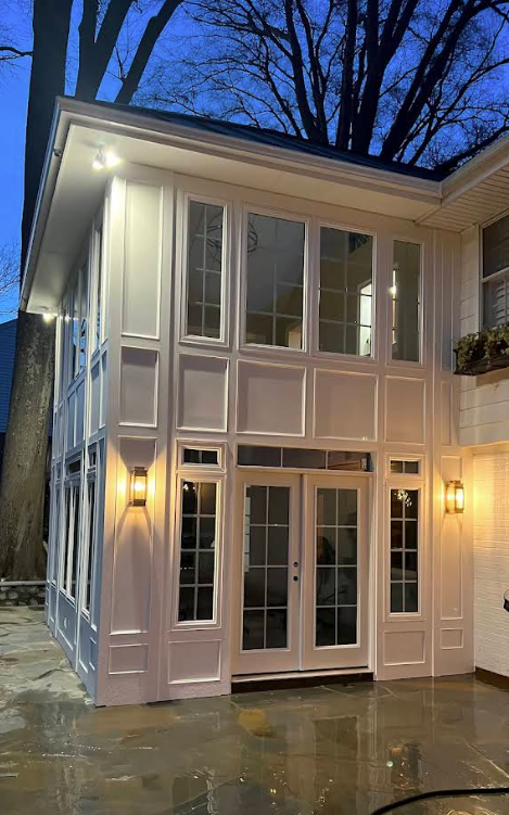 Modern two-story white house with tall windows and lit sconces at dusk, wet patio in front