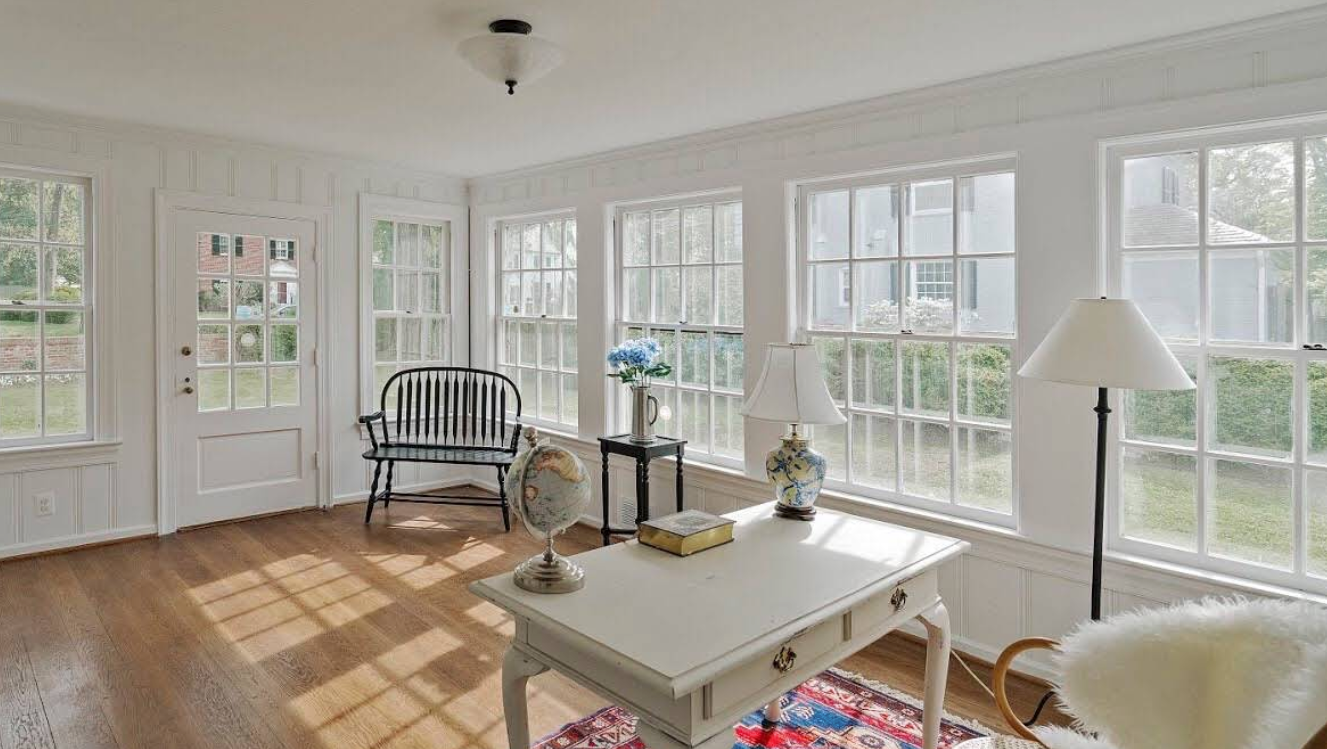 Bright sunroom with large windows, a white coffee table, chair, lamp, and wooden floor.
