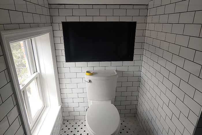Small bathroom with a toilet, wall-mounted TV, white subway tile, and a window on the left.