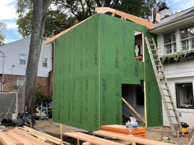 Green sheathed house addition under construction beside a ladder and lumber in a residential yard