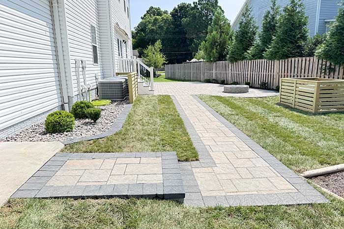Stone patio and walkway beside a white house, leading through a landscaped backyard with grass and trees.