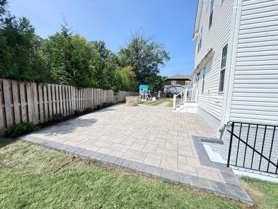 Backyard patio with stone pavers, grass edge, wooden fence, and white house exterior under a clear blue sky