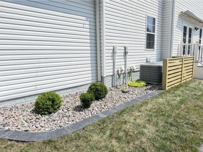 Side yard landscaping with shrubs, stones, utility units, and a beige privacy screen beside a white house.