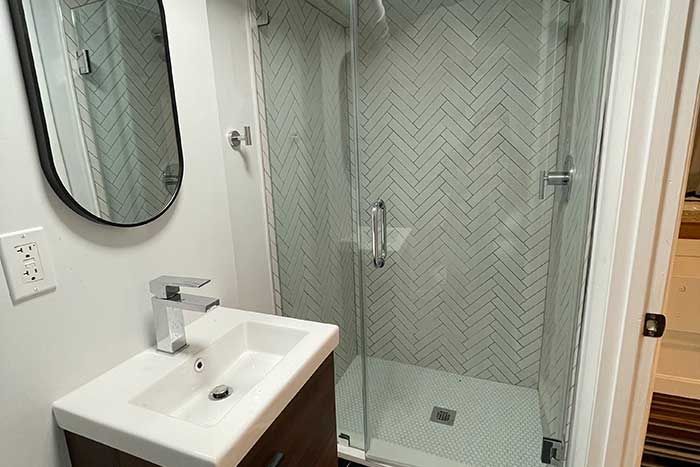 Small bathroom with pedestal sink, oval mirror, and glass shower enclosure with herringbone tile.