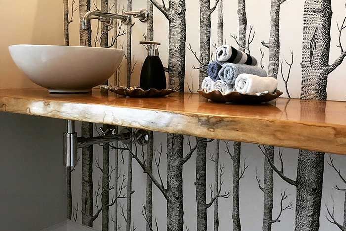 Bathroom countertop with a white bowl sink, soap dispenser, and rolled towels on a wooden shelf with tree wallpaper.