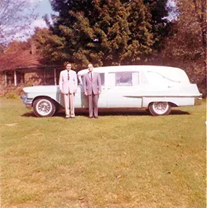 Two funeral directors standing in the distance in front of white hearse Mynatt Funeral Home, Inc.
