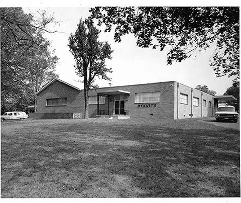 Black and white photo of Mynatt Funeral Home, Inc that reads Mynatt's with hearse on side of building