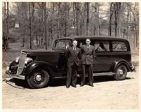 Early 1900's black and white photo of two funeral directors for Mynatt Funeral Home, Inc in front of  old hearse with grove of trees in background
