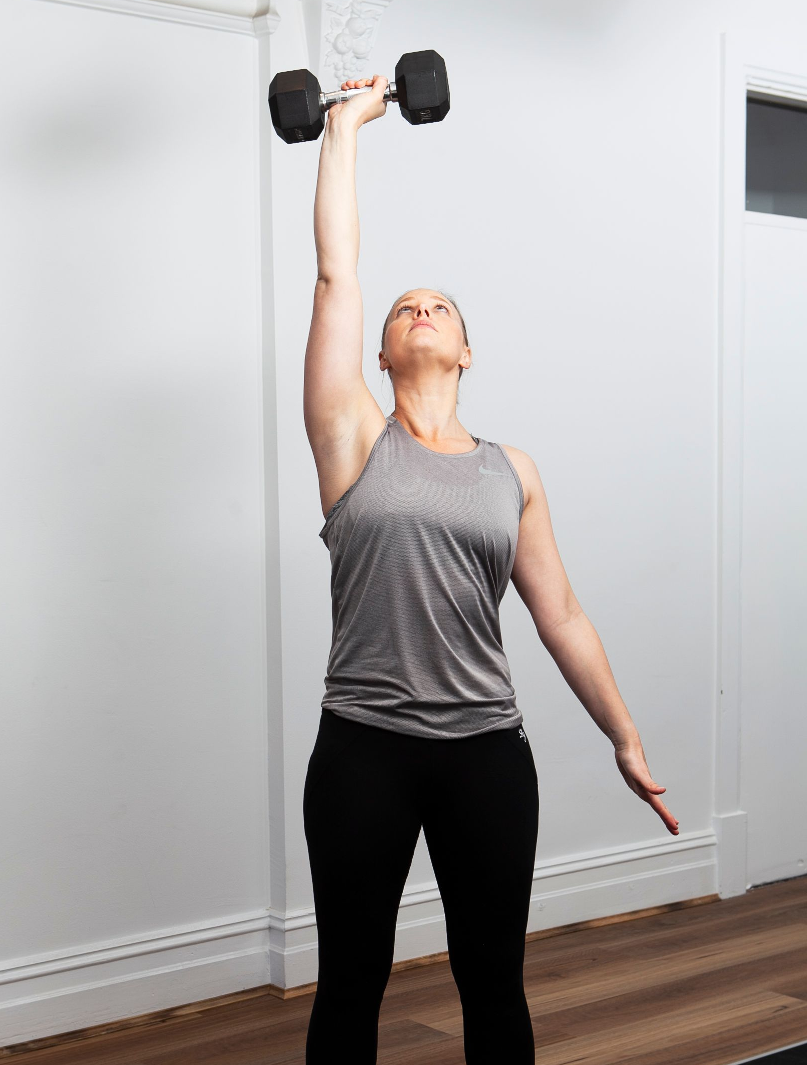 Woman in workout attire raises a dumbbell overhead in a light-filled room.