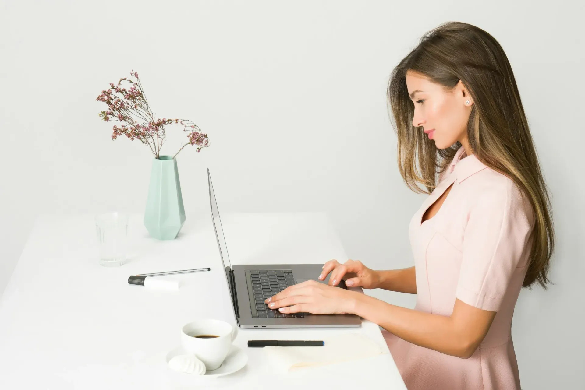 Woman typing on laptop at white desk; coffee, vase of flowers.