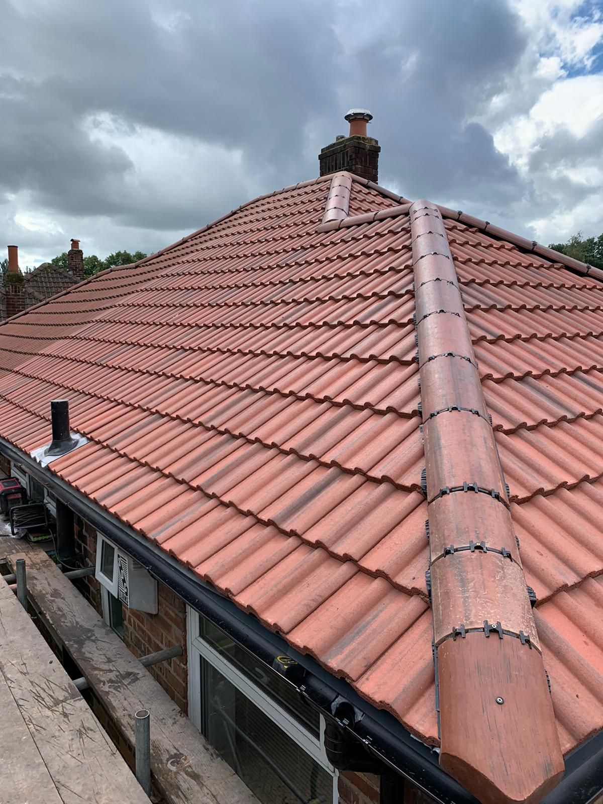 Red tiled roof with a central metal ridge and black gutters against a cloudy sky.