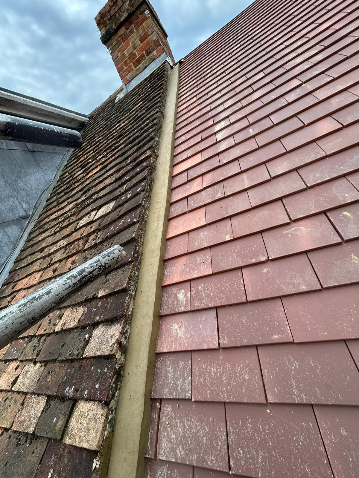 Roof with two sections of tiles, one weathered and one newer, next to a chimney and gutter.