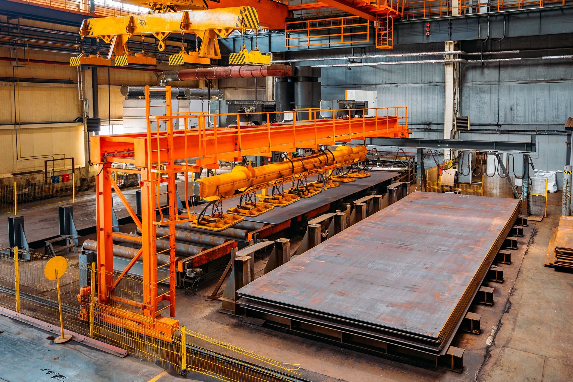 Industrial crane lifting metal sheets in a factory setting, with orange and gray machinery.