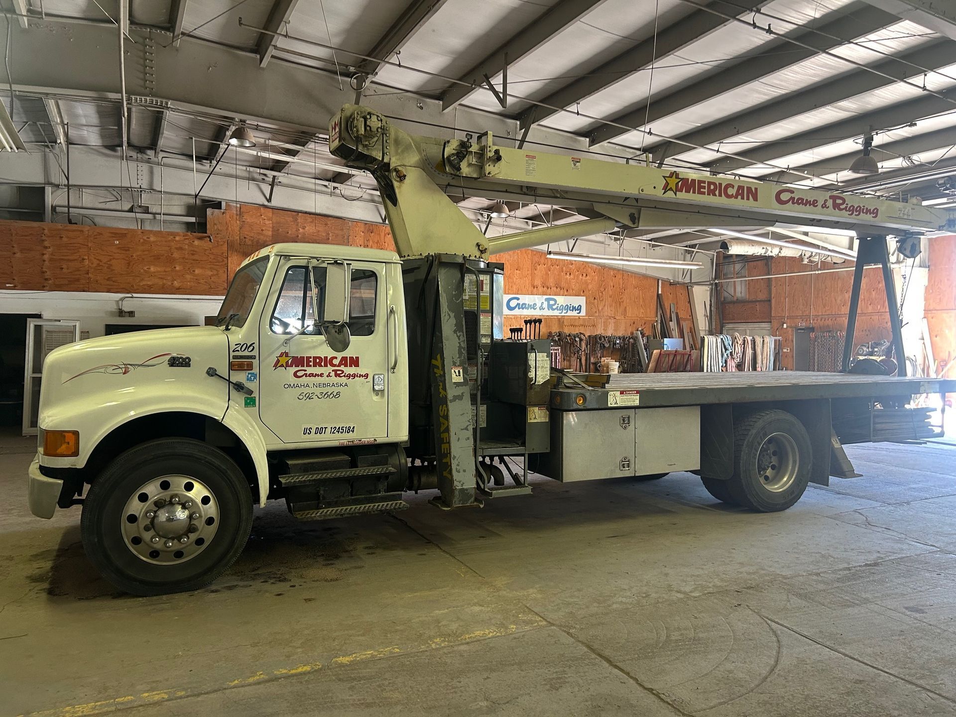 White truck with a crane arm extended over a flatbed, driving on a road. Blue sky in the background.