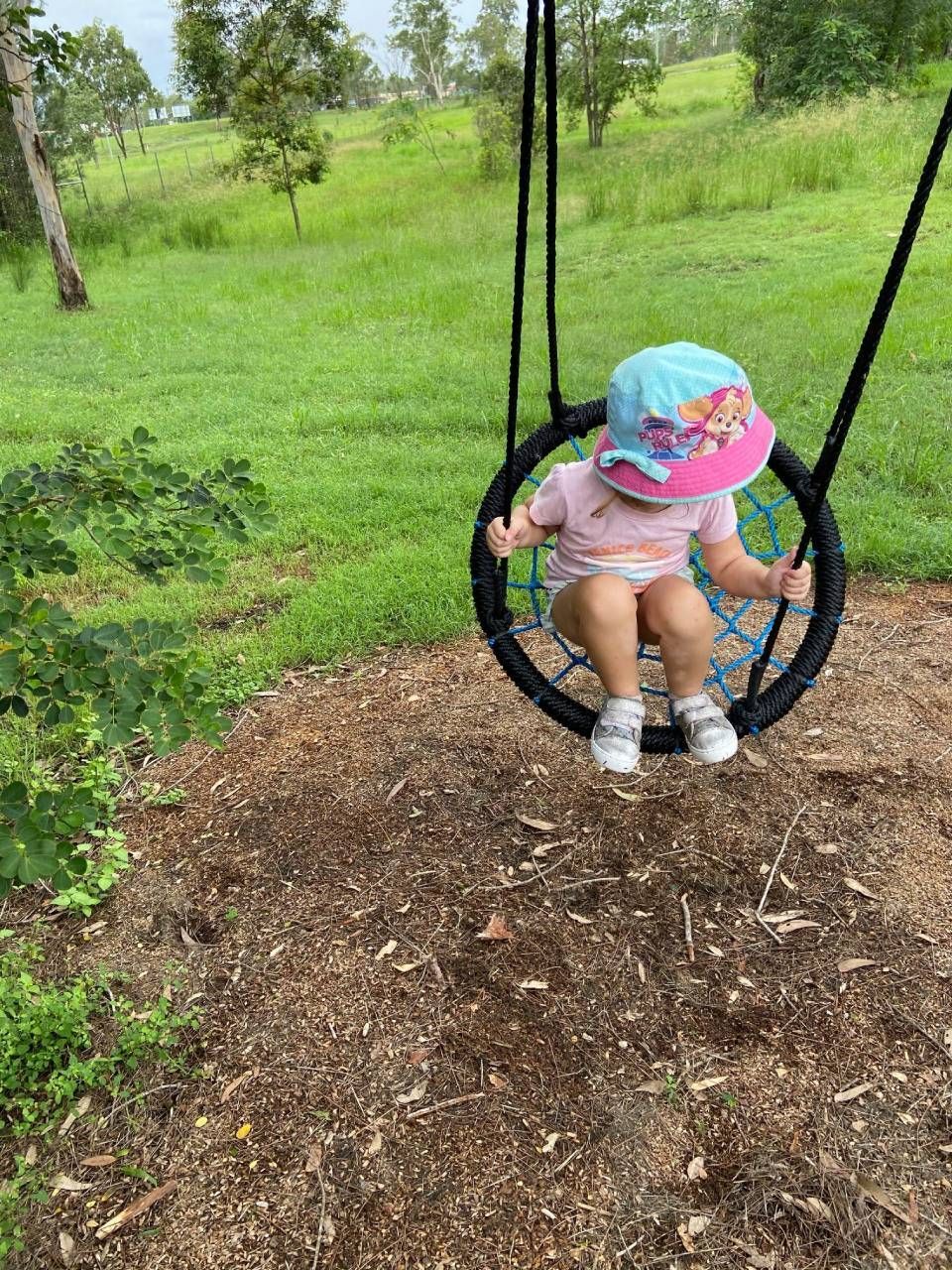 Child sitting on a black rope swing over mulch in a grassy park, wearing a pink hat and blue outfit.