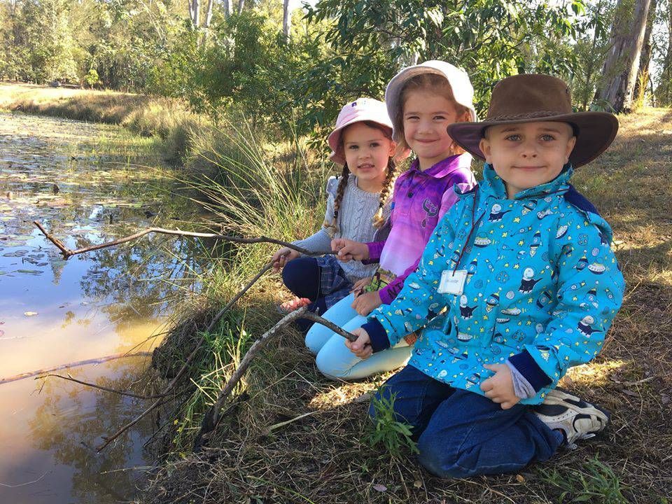 Three children in hats sit beside a creek, fishing in a sunny wooded area.