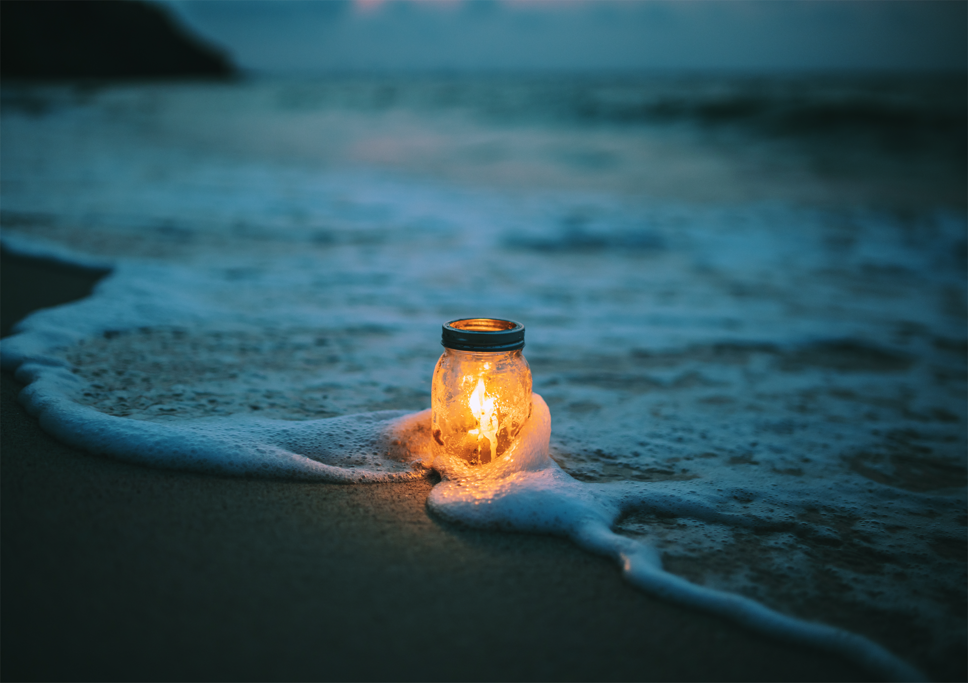 Glowing jar candle on wet sand by the shoreline at dusk