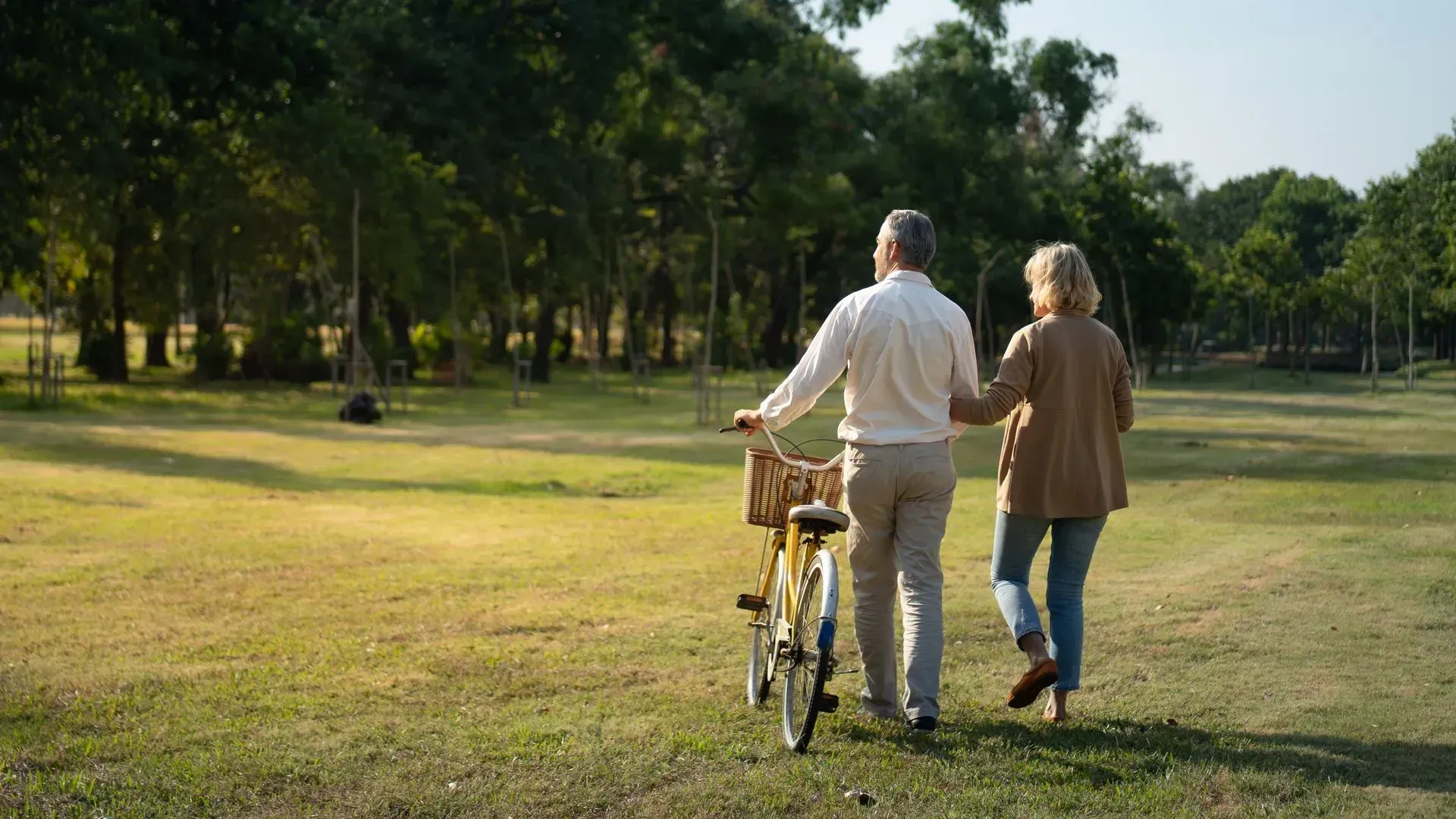 Two people walking a bicycle across a sunny grassy park near trees