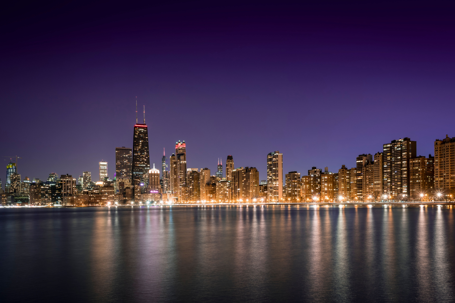 City skyline at night with lit skyscrapers reflected on calm water under a purple sky