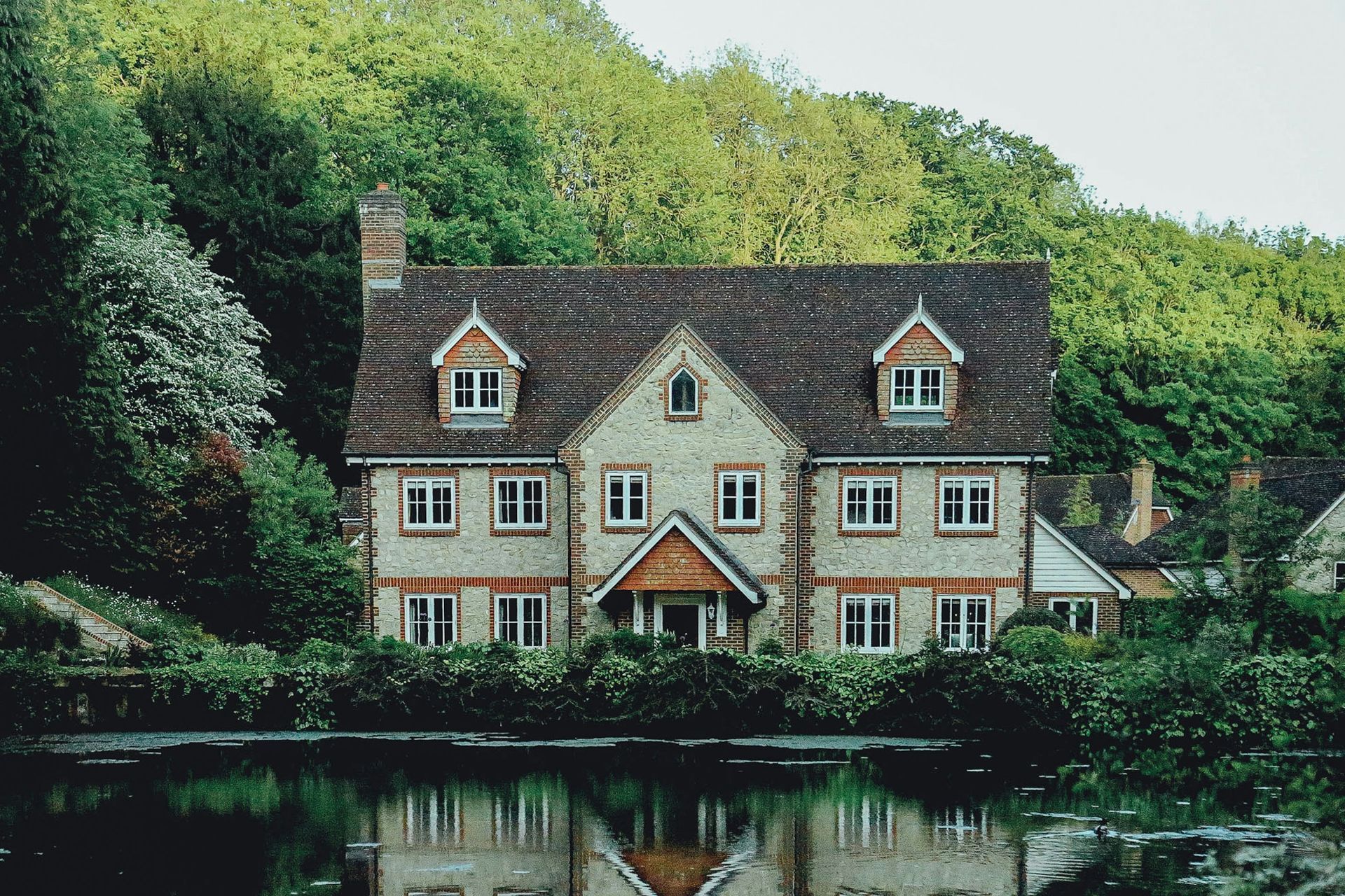 Charming white cottage by a pond, surrounded by lush green trees and reflected in the water.
