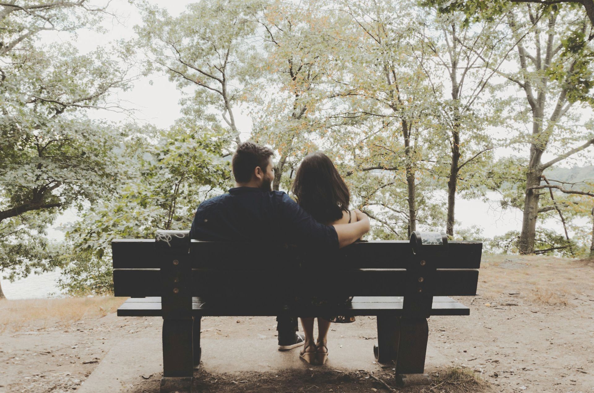 Two people sit on a bench under trees, looking out over a quiet lakeside path.