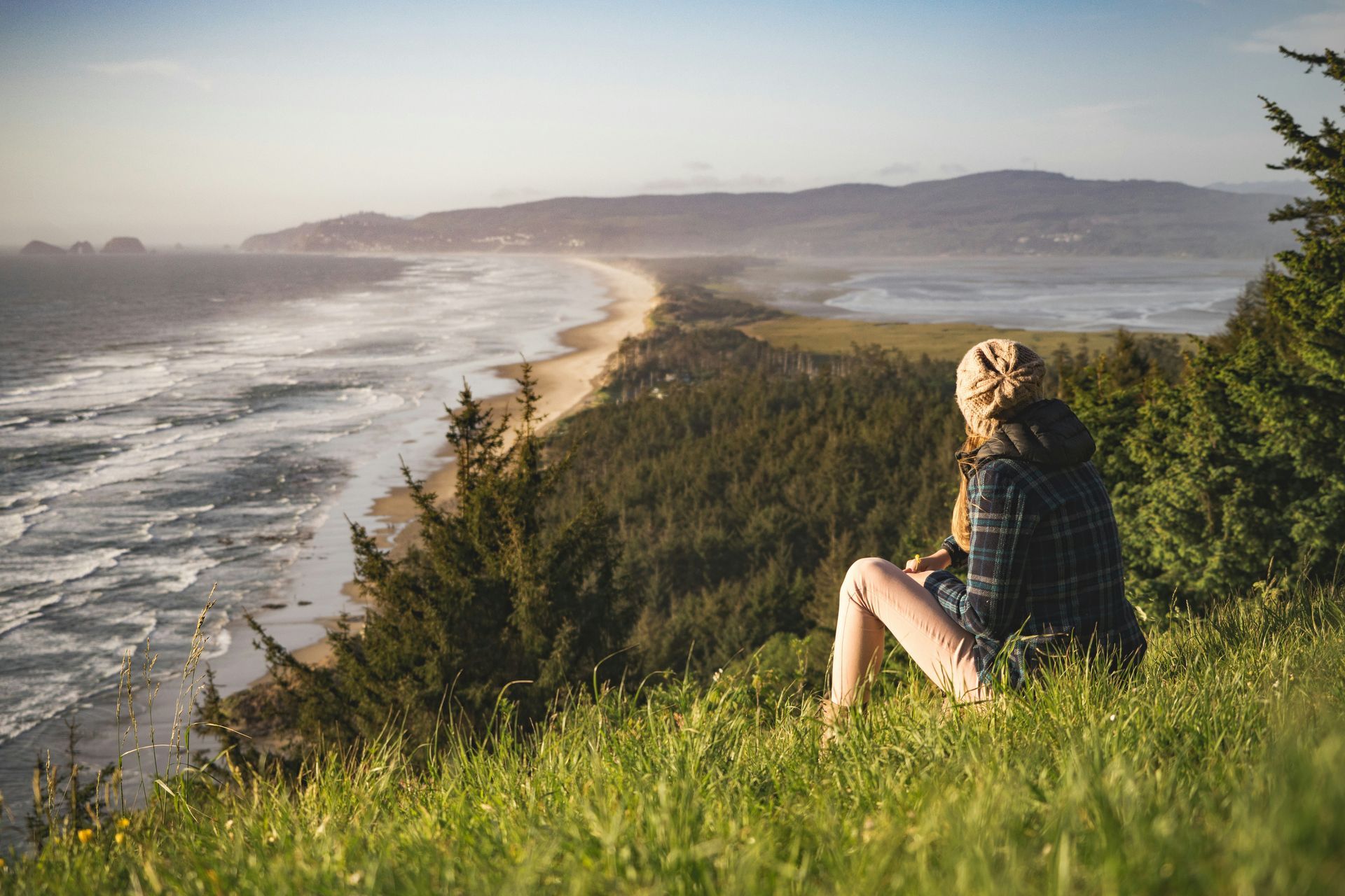 Person sits on a grassy cliff overlooking a beach and ocean at sunset.