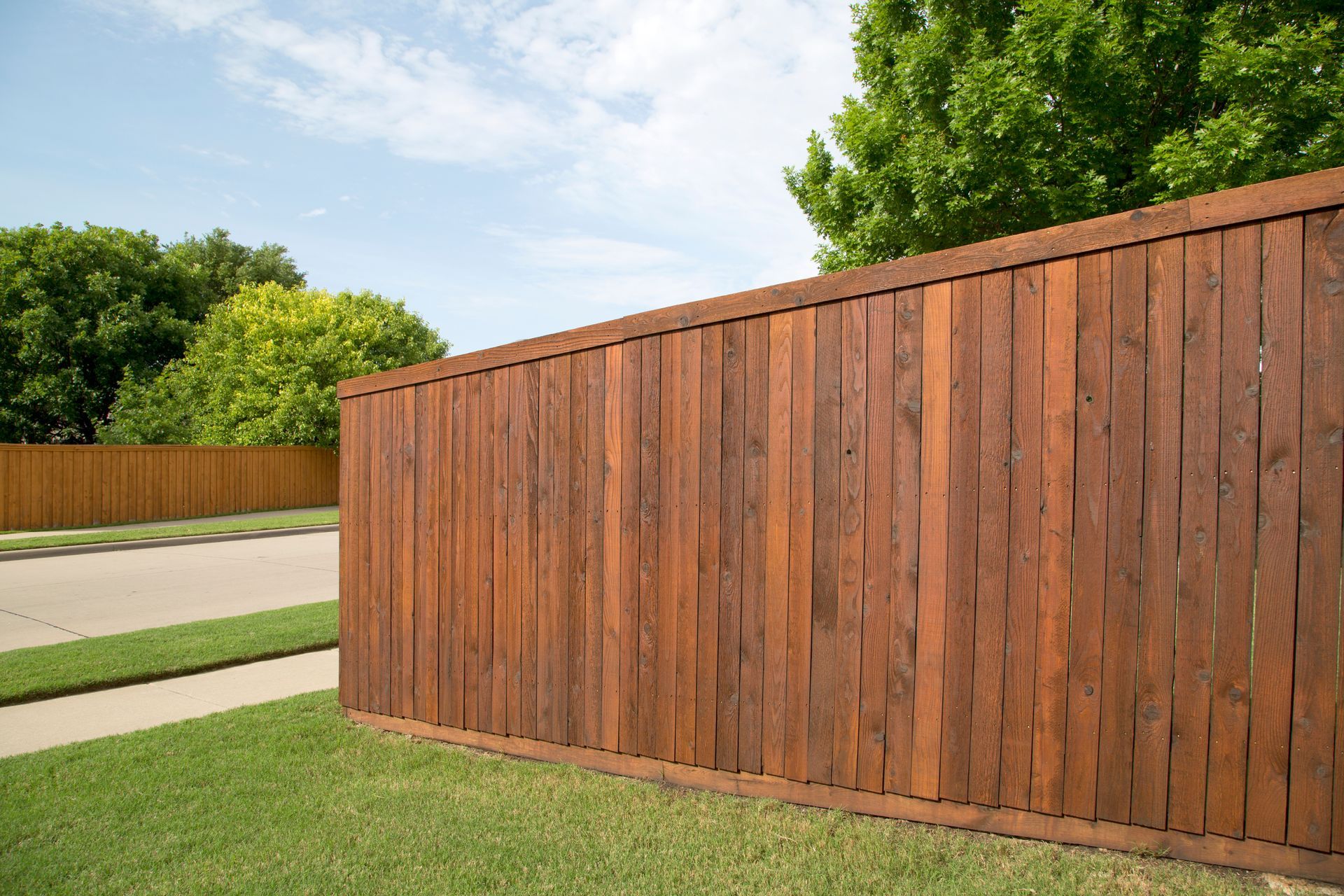 Brown wooden privacy fence along a green lawn and a street, with trees in the background.