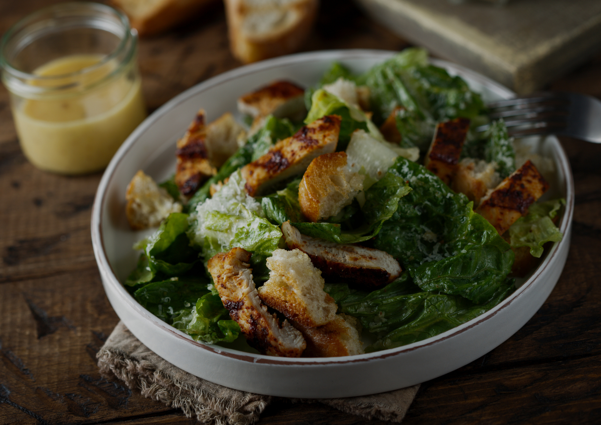 Salade César avec croûtons et copeaux de fromage, servie dans un saladier blanc sur une table rustique, avec la vinaigrette.