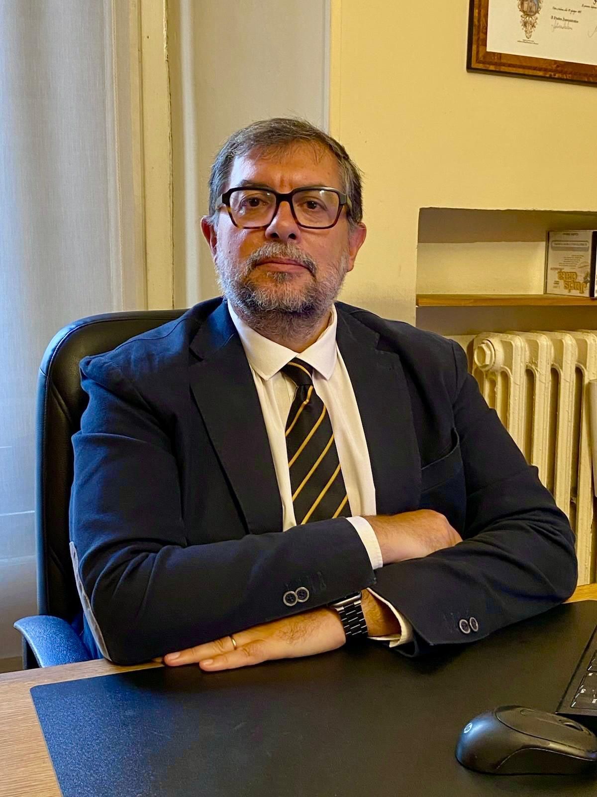 Un uomo in abito scuro e occhiali è seduto a una scrivania in ufficio, con le braccia incrociate.