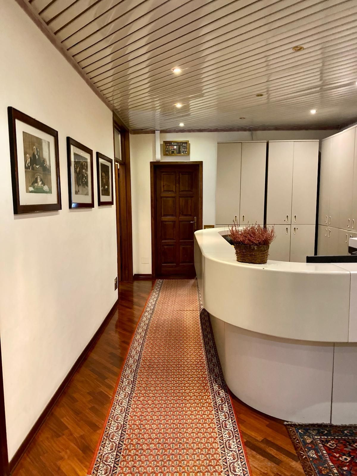 Lungo corridoio con quadri incorniciati, pavimento piastrellato con motivi decorativi e un bancone reception bianco e curvo.