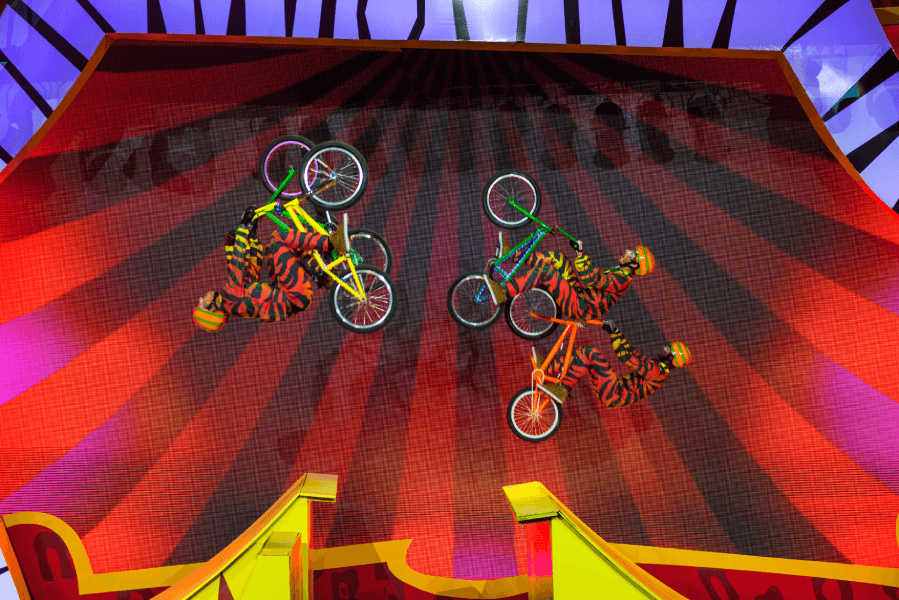 Dois ciclistas de BMX com roupas estampadas realizam manobras no ar em um palco listrado em vermelho e preto.
