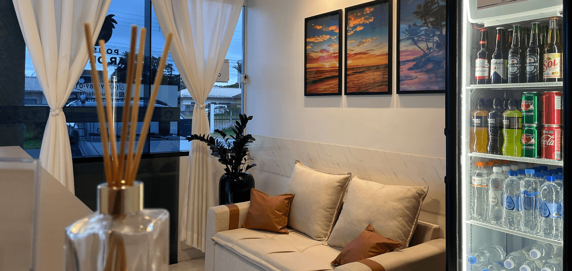 Uma área de estar aconchegante com sofá, almofadas decorativas, obras de arte na parede e um frigobar com porta de vidro abastecido com bebidas.