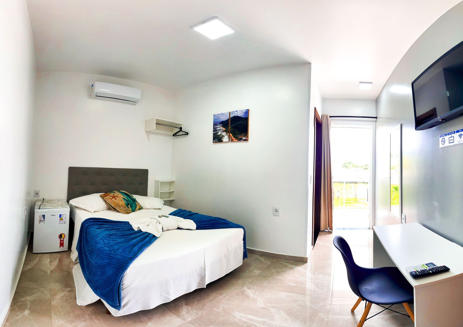 Um quarto de hotel moderno com cama, manta azul, frigobar, escrivaninha com cadeira, ar-condicionado e porta para varanda.