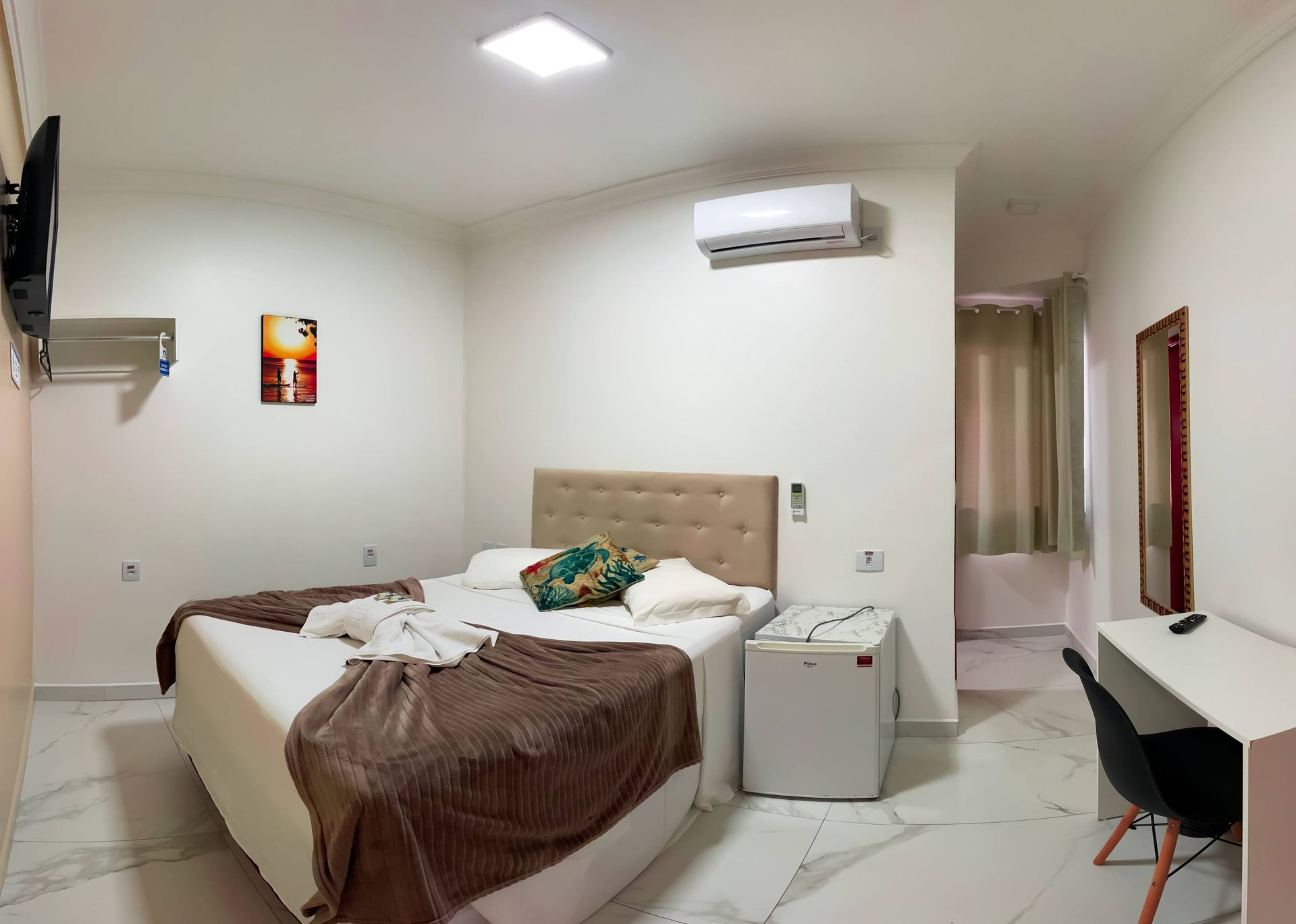 Um quarto de hotel impecável com uma cama de casal, uma colcha marrom, uma pequena geladeira branca, um ar-condicionado de parede e uma escrivaninha.