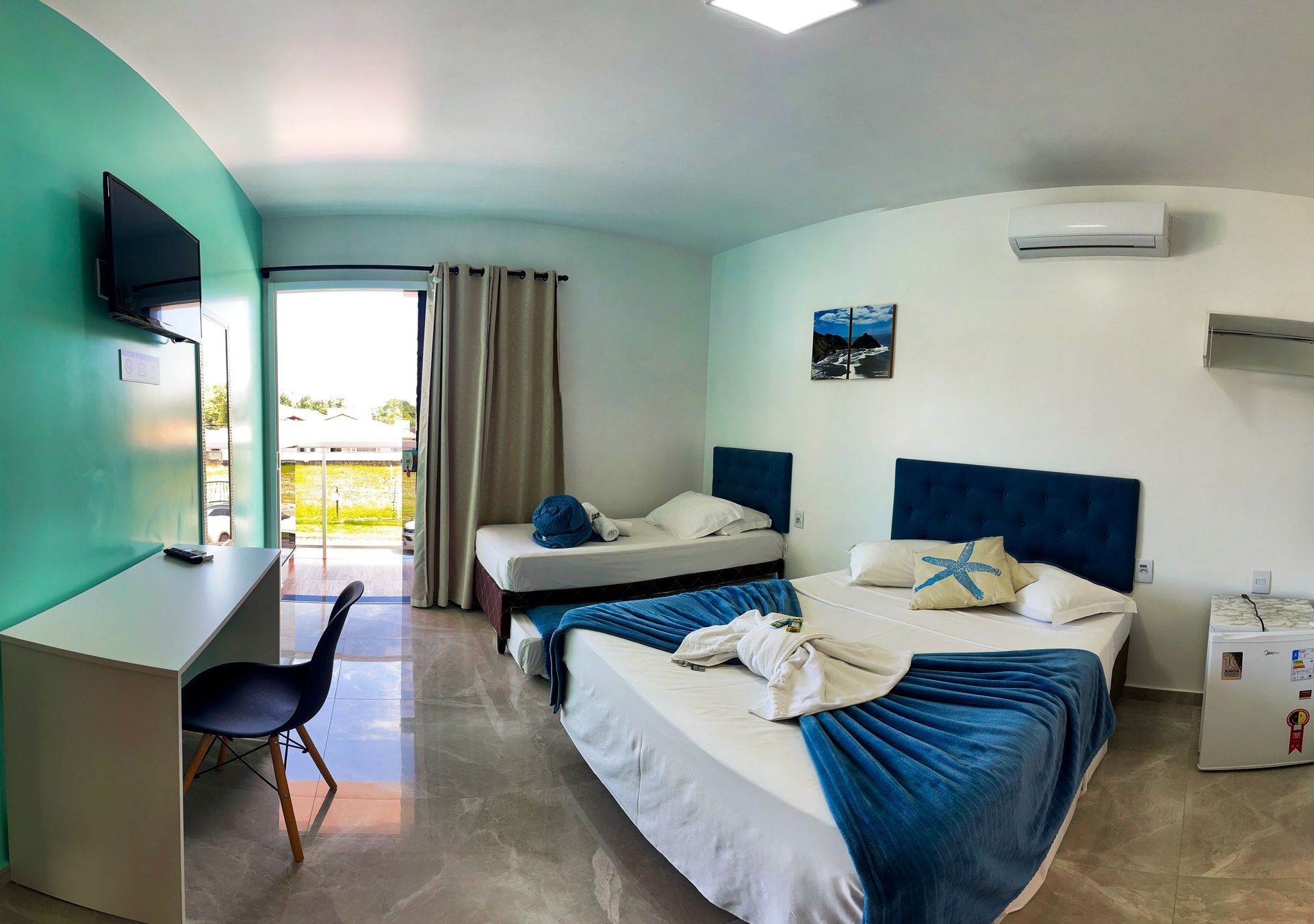 Um quarto de hotel com uma cama de casal, uma cama de solteiro, uma escrivaninha com cadeira e uma porta de correr que dá para uma área ensolarada.