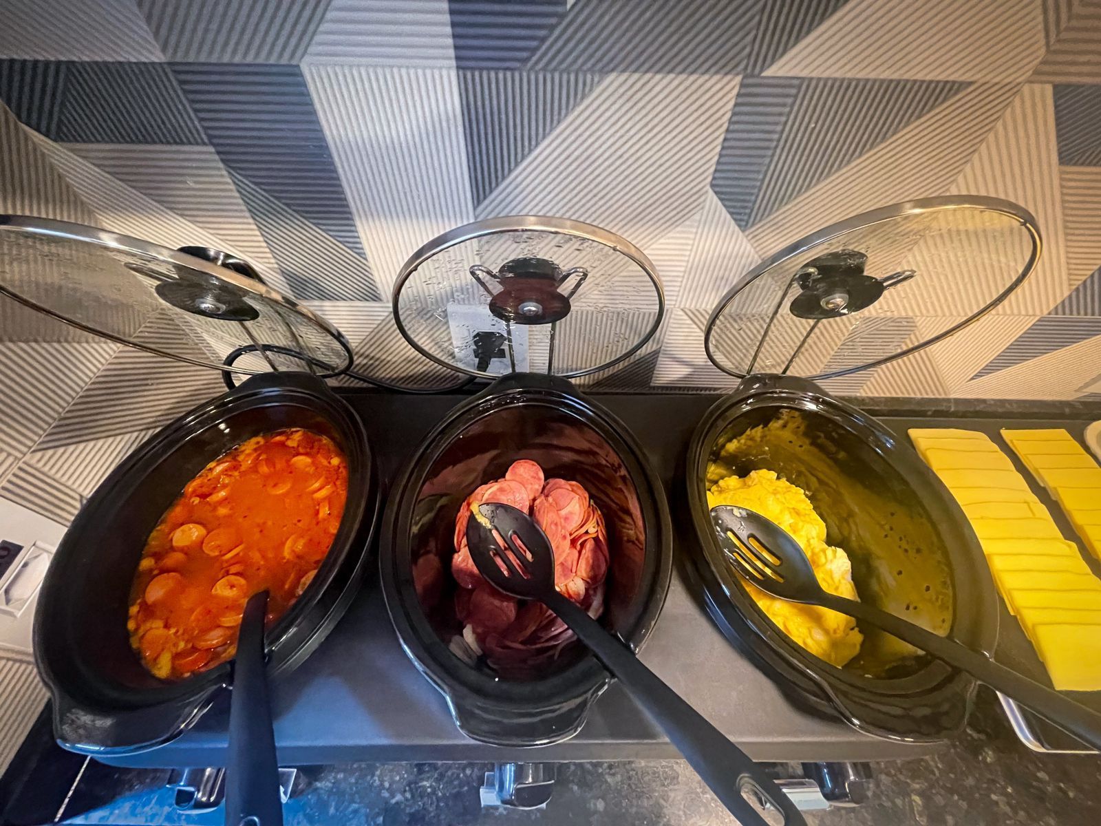 Três panelas elétricas de cozimento lento em cima da bancada: uma com feijão cozido, uma com linguiça fatiada, uma com ovos mexidos e uma com torradas.