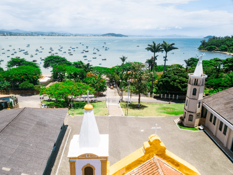 Vista elevada do pátio de uma igreja costeira, com sua arquitetura histórica, debruçada sobre uma baía repleta de pequenos barcos.