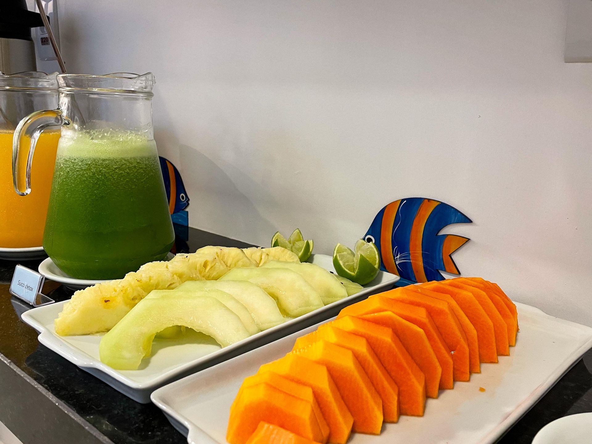 Um buffet de café da manhã com abacaxi, melão e mamão fatiados, acompanhados de jarras de suco verde e laranja.