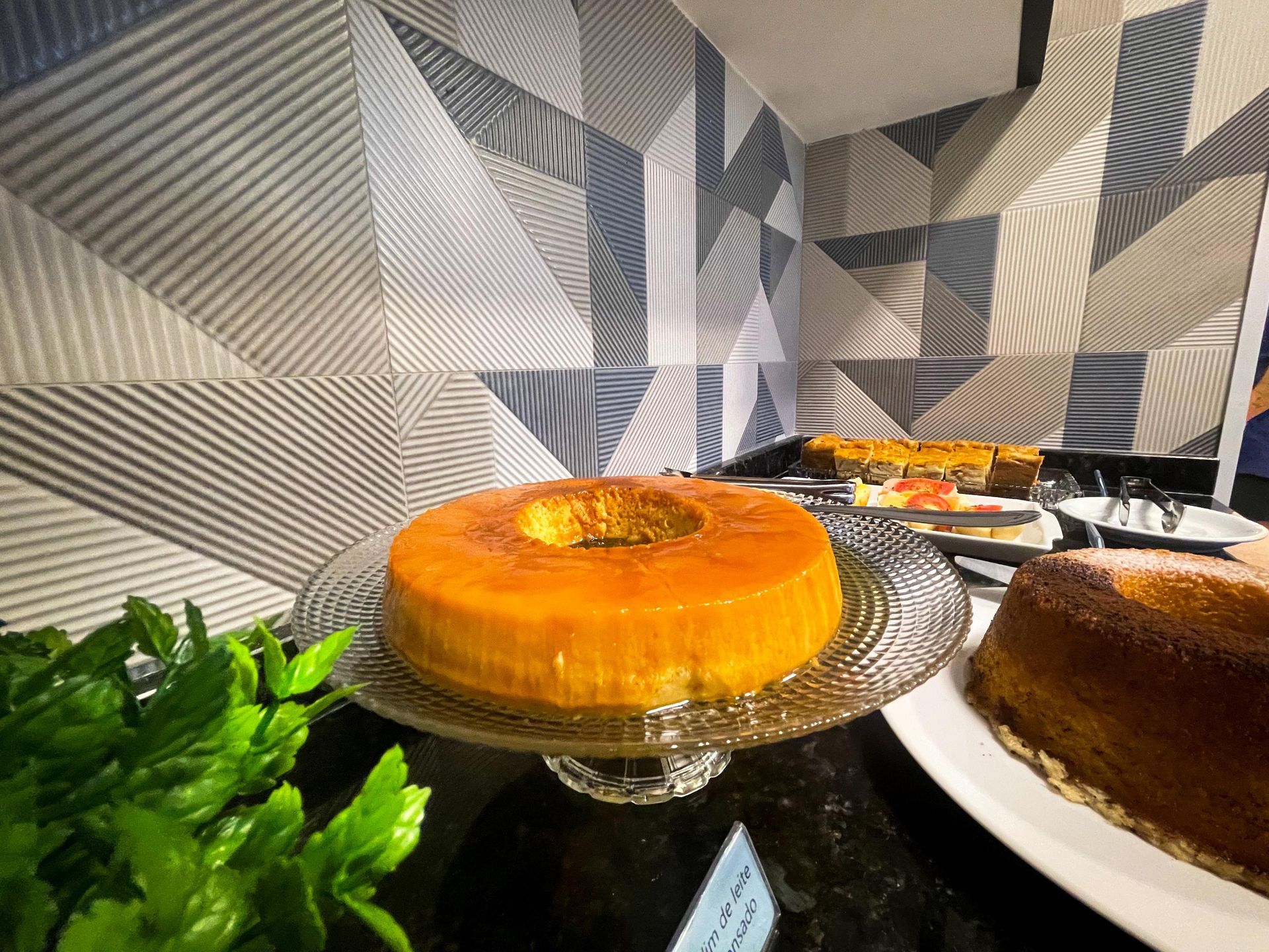 Um pudim laranja vibrante repousa sobre um pedestal de vidro ao lado de outro bolo em uma mesa de buffet com paredes revestidas de azulejos geométricos.